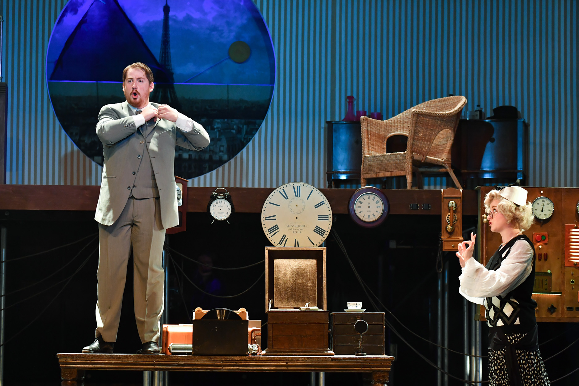 A man and women, wearing period costume, the man standing on a woodent able, surrounded with various different clocks, with the Eiffel Tower behind them in a window.